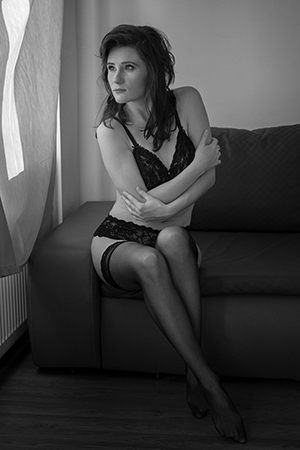 A woman in black lingerie and stockings posing by the window sitting on a dark gray sofa.