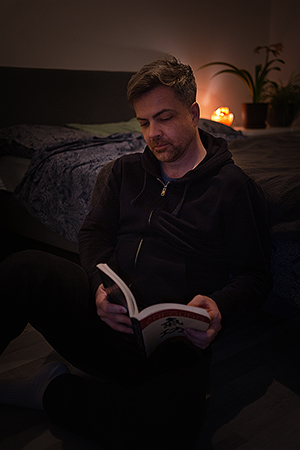 A young man in a dark room, sitting on the floor by a bed, reading a book.