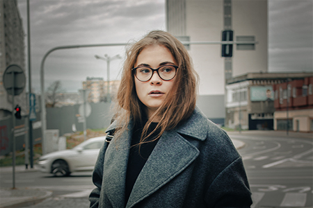 A city street portrait of a brunette wearing reader glasses and a gray coat in a gray urban scenery a cloudy day.