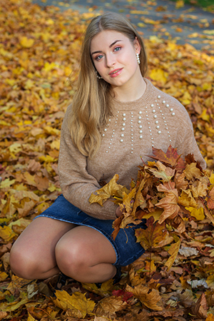 A blonde girl with blue eyes posing around autumn leaves.