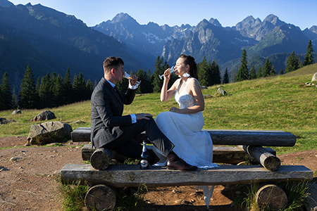 Bride and groom sitting on a wooden table, at Tatra mountains, enjoying a glass of martini champagne.