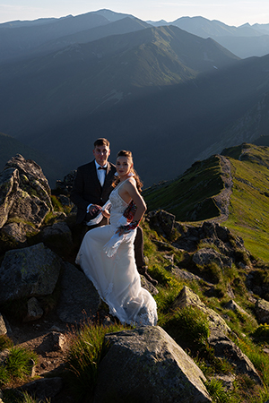 Bride and groom are posing and smiling infront of a green mountain hillside.