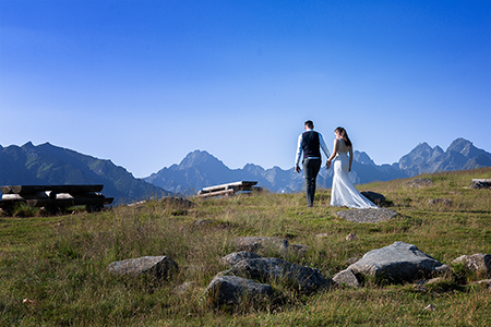 Groom holding bride by hand, walking on the grass among rocks and wooden tables, with high rocky montains on the distance.