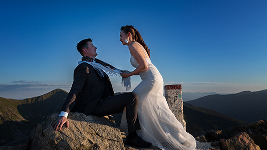 Bride is pulling groom with a scarf around his neck, sitting to the rocks and a milestone at Tatra mountains in Poland.