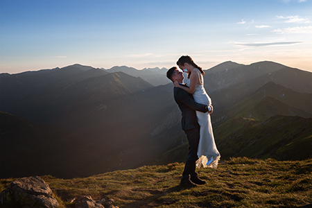 Groom lifts up in his hug the bride, in a treeless mountainscape, a sunny evening.