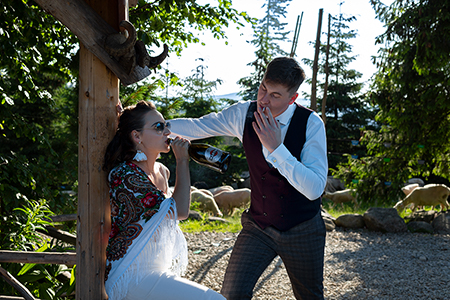 Groom is smoking and bride drinking from bottle in a wooden house with sheeps eating grass in background.