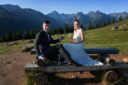 Wedding couple sitting on a wooden table raising their champagne glasses with mountain hills in the distant background.