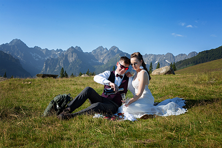 Groom and bride with a green backpack and sunglasses sitting on the grass at a mountain hill. Groom is pouring champagne to bride's glass.